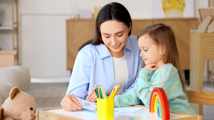 A smiling female psychologist sits at a small table with a young girl during an art therapy session, surrounded by colored pencils, drawings, and a teddy bear.