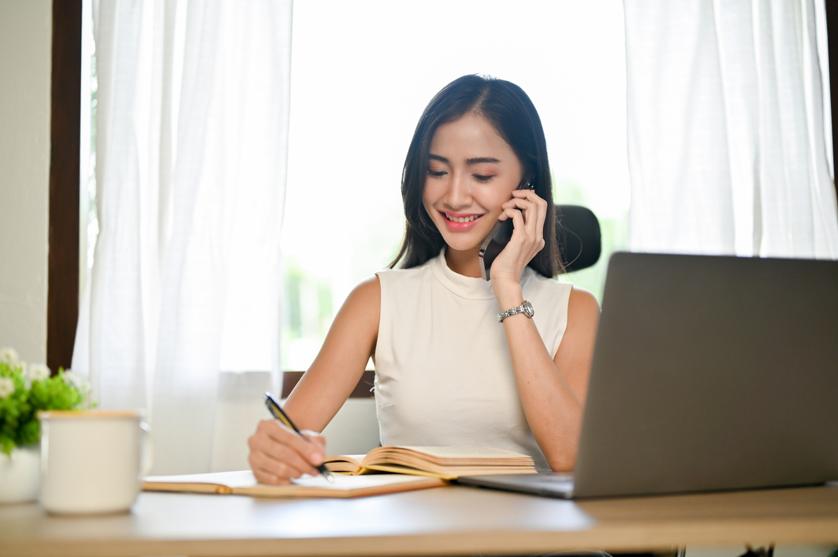 Young Woman Sits in Front of a Laptop Holding a Cellphone to Her Ear