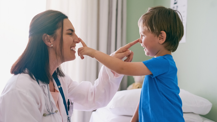 A female pediatrician in a white lab coat playfully poking the nose of a young boy during a medical exam in a bright clinic.