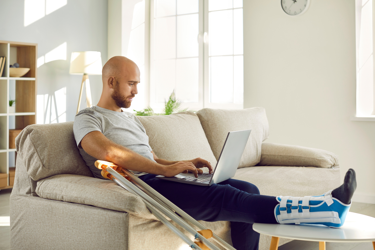 Male With Injured Leg In Bandage Sits on Couch With Laptop