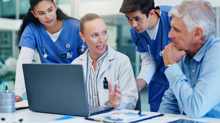 A diverse group of healthcare professionals, including doctors and a surgeon, gathered around a laptop in a hospital setting for a collaborative discussion.