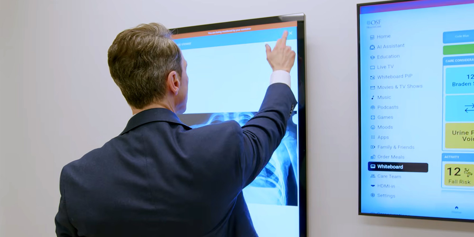 A man in a professional suit, viewed from behind, interacting with a large touchscreen display showing a digital interface with a blue header and white content area.