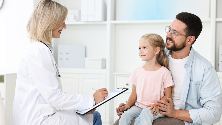 A blonde female doctor in a white coat holding a clipboard while speaking with a young girl and a man with glasses in a modern hospital setting.