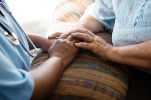 Close-up of a nurse in blue scrubs gently holding an elderly patient's hands on a patterned cushion, offering comfort and support.