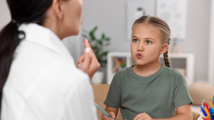 A female speech therapist in a white coat working with a young girl at a desk, helping her with reading or writing exercises from an open book.