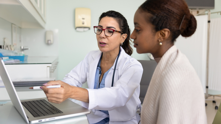 Female doctor showing examination result to young woman patient, pointing at laptop screen, explaining electronic medical report.