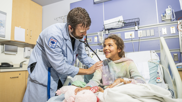 A male doctor using a stethoscope to listen to a young female patient's heart. The girl is in a hospital bed holding a stuffed animal and smiling.