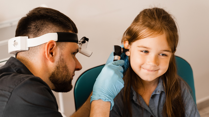 A male pediatric otolaryngologist wearing a head mirror and blue gloves uses an otoscope to examine the ear of a smiling young girl in a clinic setting.