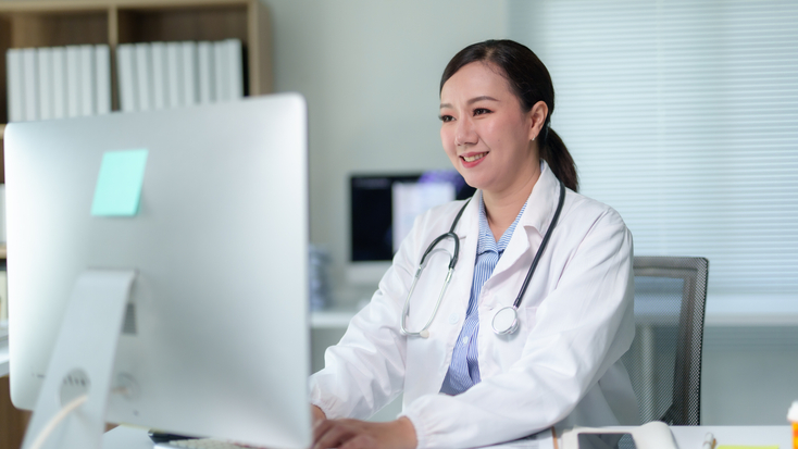 A female doctor wearing a lab coat and stethoscope working on a desktop computer in a medical office.