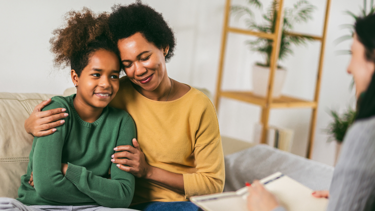 A supportive mother and her teenage daughter sitting on a couch together during a family counseling or therapy session.