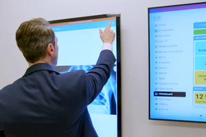 A man in a professional suit, viewed from behind, interacting with a large touchscreen display showing a digital interface with a blue header and white content area.