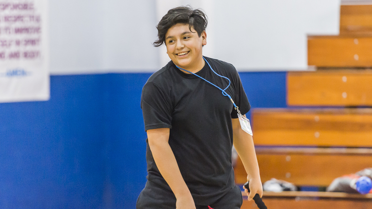 A young person in a black t-shirt smiling while participating in a Healthy Kid's University program at a YMCA, standing in front of gymnasium bleachers during a physical activity session.