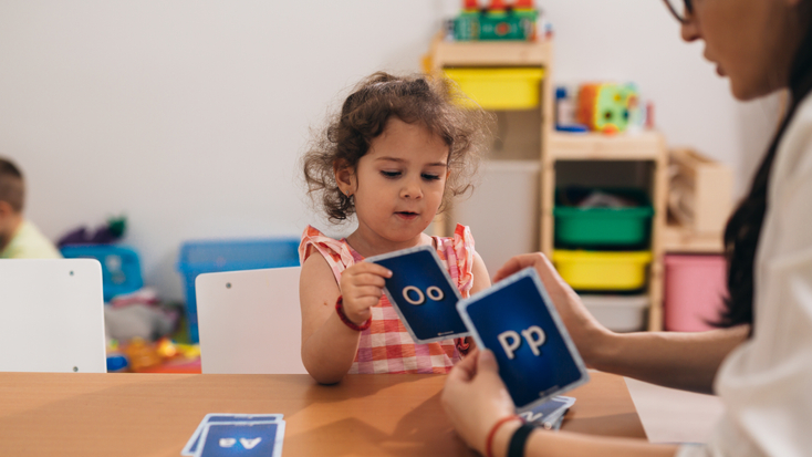A young girl in a plaid shirt sits at a desk, looking at flashcards while a speech therapist in a white coat guides her on pronunciation.