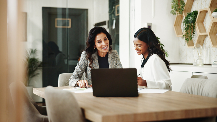 Two smiling businesswomen sitting at a wooden desk and collaborating over a laptop in a modern office.