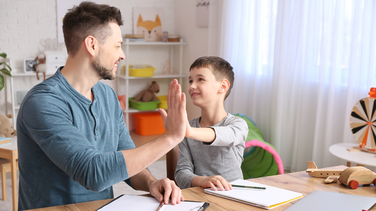 A male psychologist sits at a desk and gives a high-five to a smiling young boy during a session in a bright office filled with toys and learning materials.