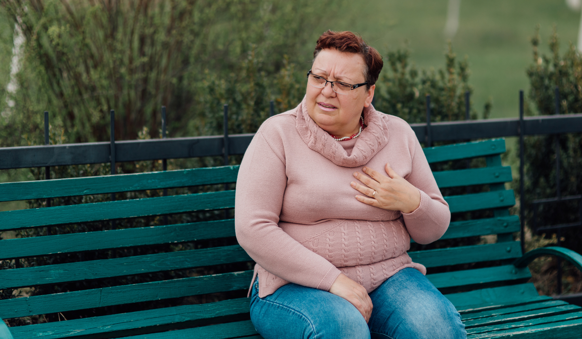 woman sits on a park bench with a pained look and a hand over her heart