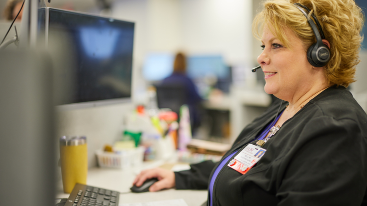 A blonde female healthcare professional wearing a telephone headset smiles while working at her computer workstation in an office environment.