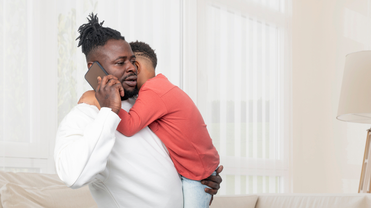 A father warmly embracing his young child in a brightly lit living room.