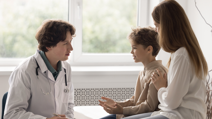 A young male pediatrician in a white coat sitting and talking to a young boy patient who is sitting with his mother during a medical checkup in a clinic.