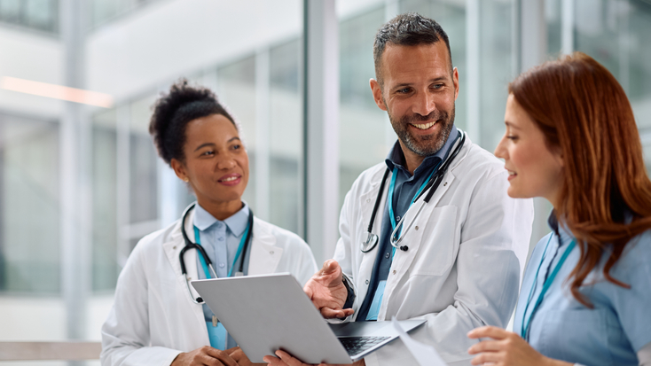 Three medical experts—a male doctor in the center, flanked by two female colleagues—working together on a laptop in a bright, contemporary clinical setting.