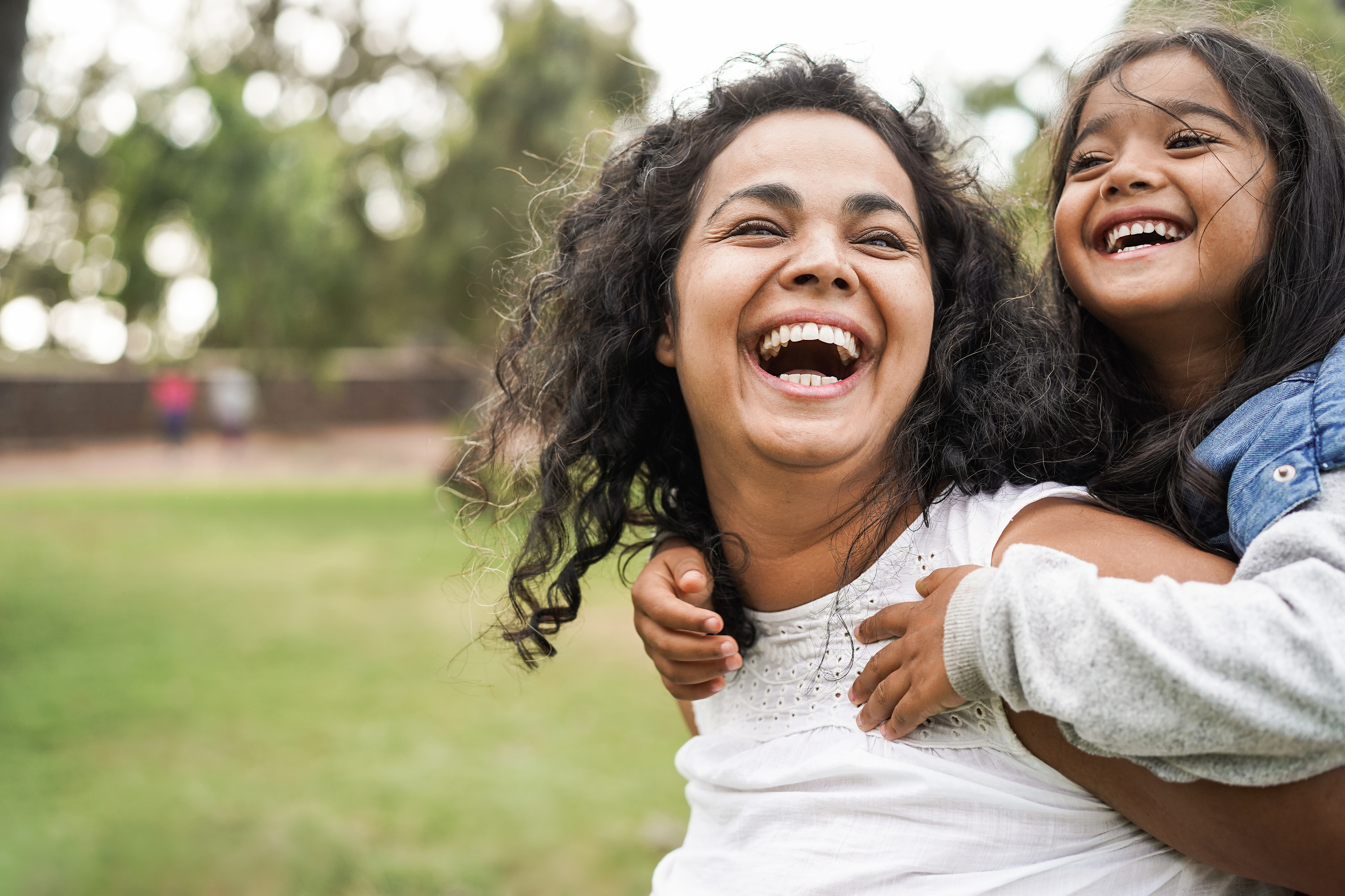 A woman smiling with a young girl on her back, both laughing in an outdoor park setting.