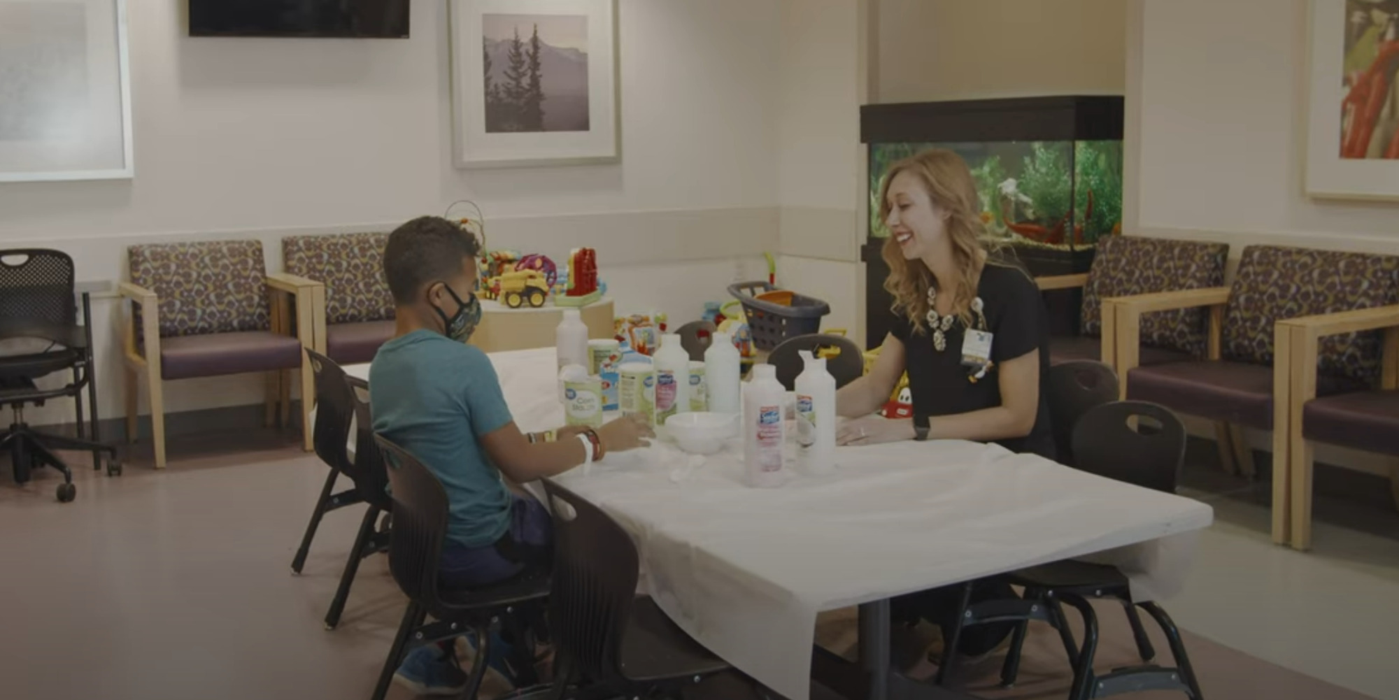 A child wearing a mask sits at a table with an adult in a playroom, surrounded by toys and an aquarium in the background.
