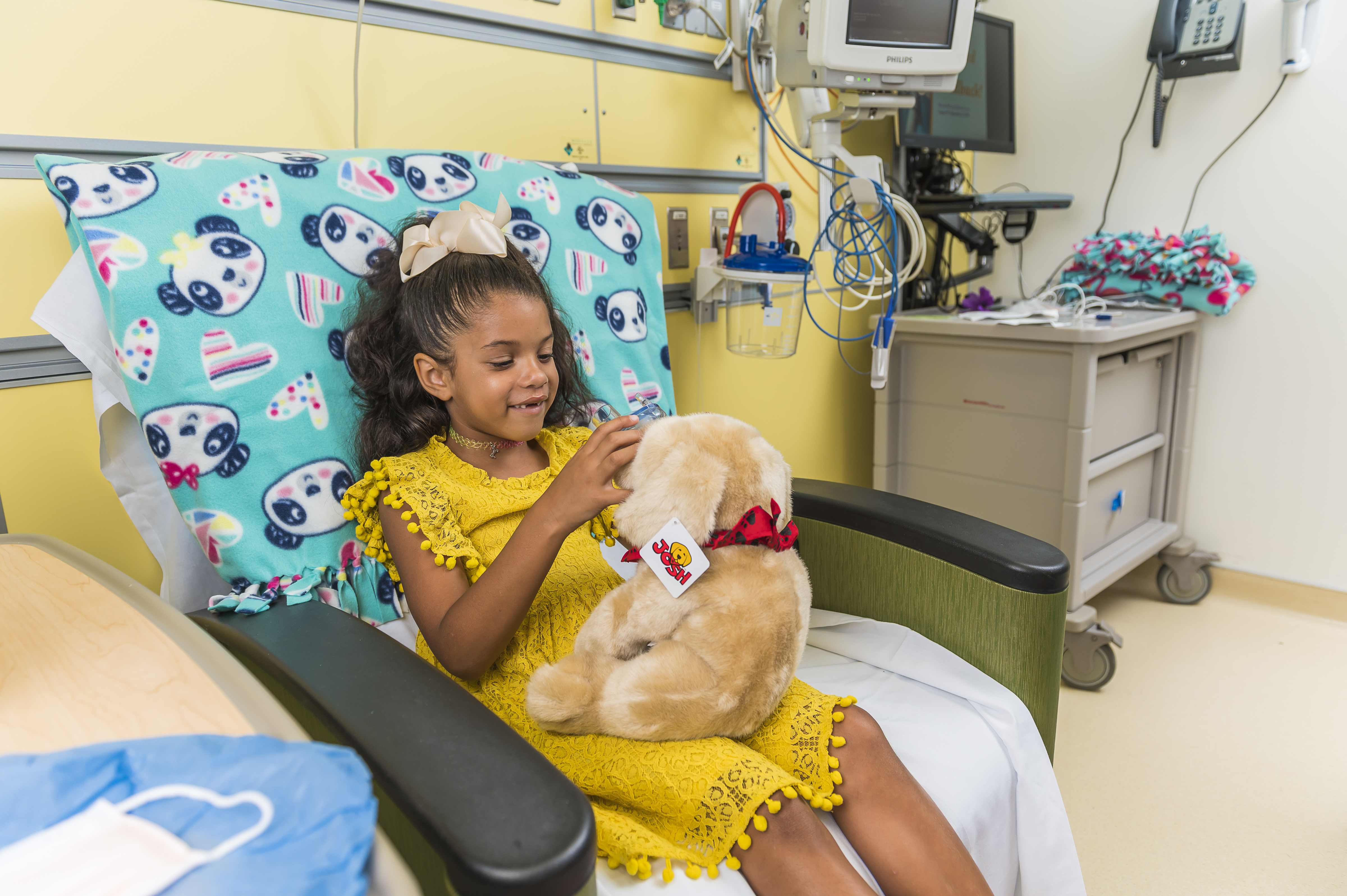 A young girl wearing a yellow dress holds a plush toy while seated in a hospital room, next to medical equipment and a colorful blanket.