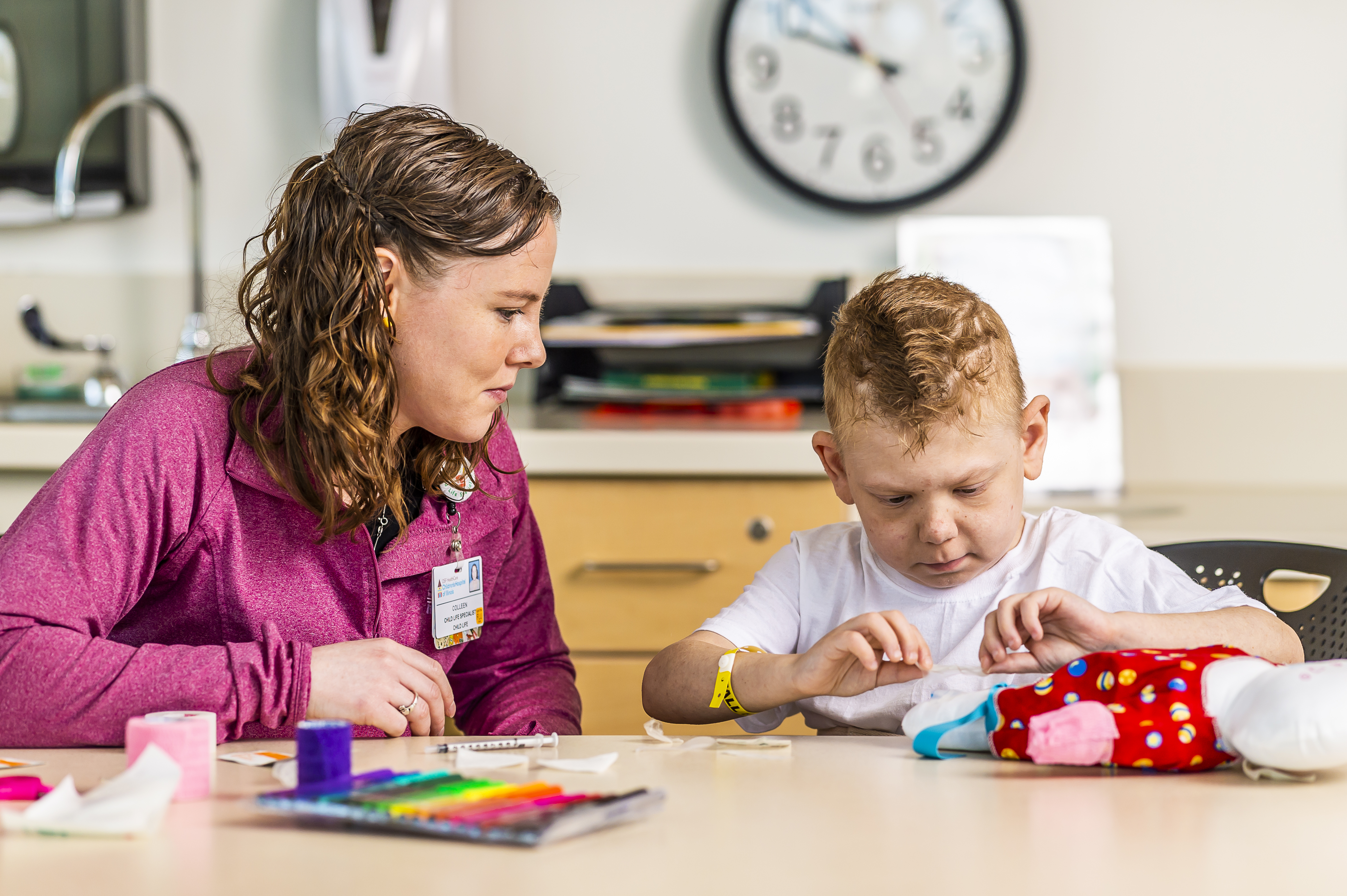 A caregiver and a child engaging in an activity at a table with craft supplies in a brightly lit room.
