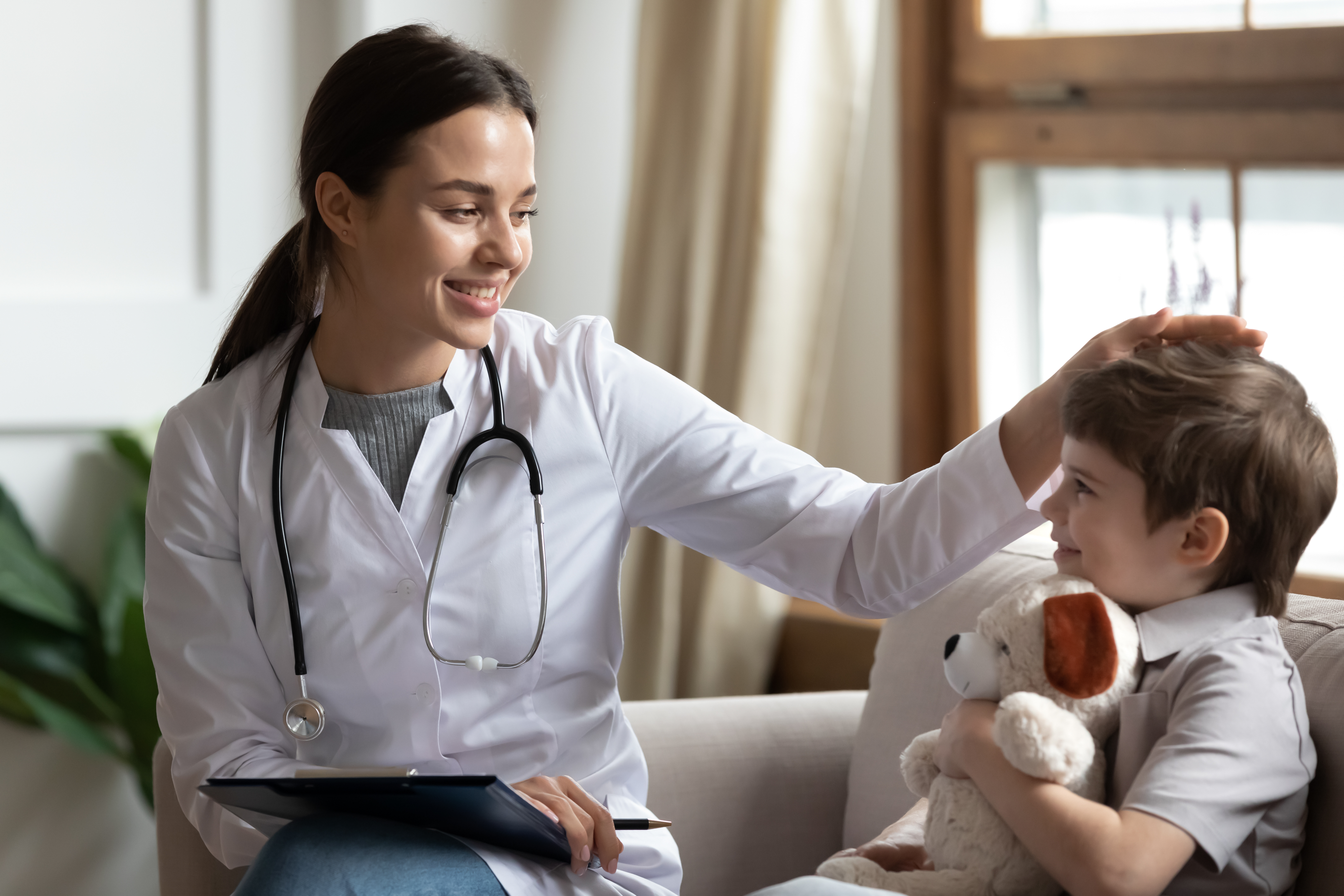 A female doctor smiling warmly while placing her hand gently on a young boy's head. The boy is holding a plush toy.