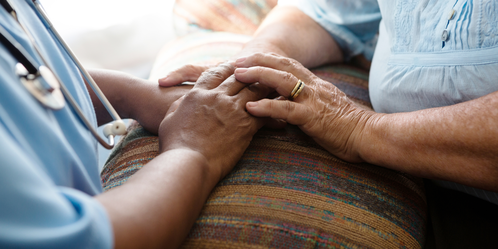 Close-up of a nurse in blue scrubs gently holding an elderly patient's hands on a patterned cushion, offering comfort and support.