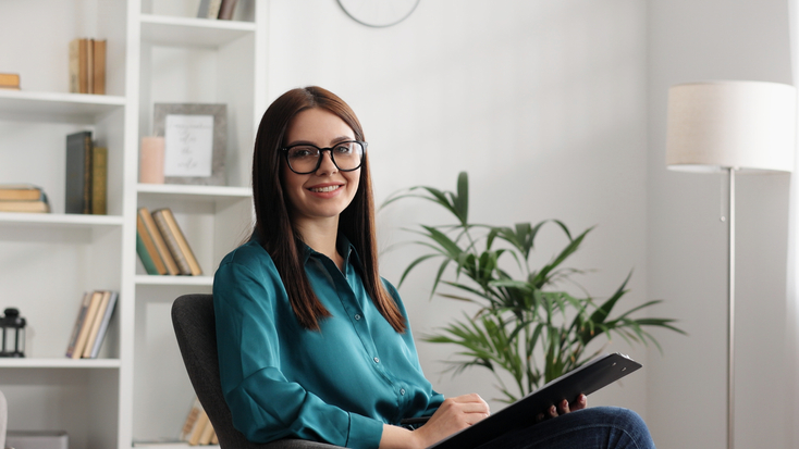 A smiling mental health professional sitting in a modern therapy office, ready for a client consultation.