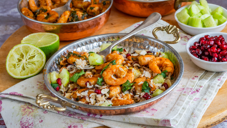 Tandoori prawns served over a bed of wild rice with fresh herbs and pomegranate seeds in a copper bowl, next to a halved lime.