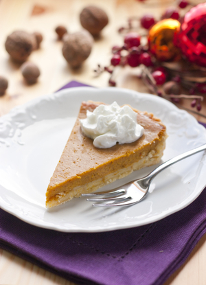 A slice of pumpkin pie topped with a dollop of whipped cream on a white plate, set against a festive holiday background with red berries and ornaments.